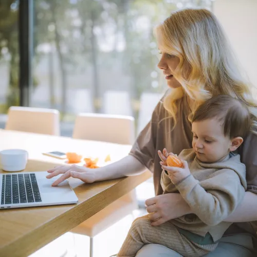 Mom sits down to work on her computer with her child eats a snack while sitting on her lap.