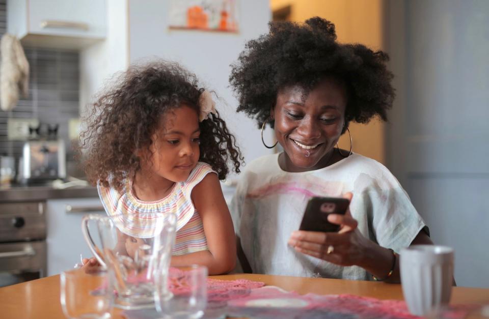 A mother smiles while looking at her cell phone as a young girl looks over her shoulder.