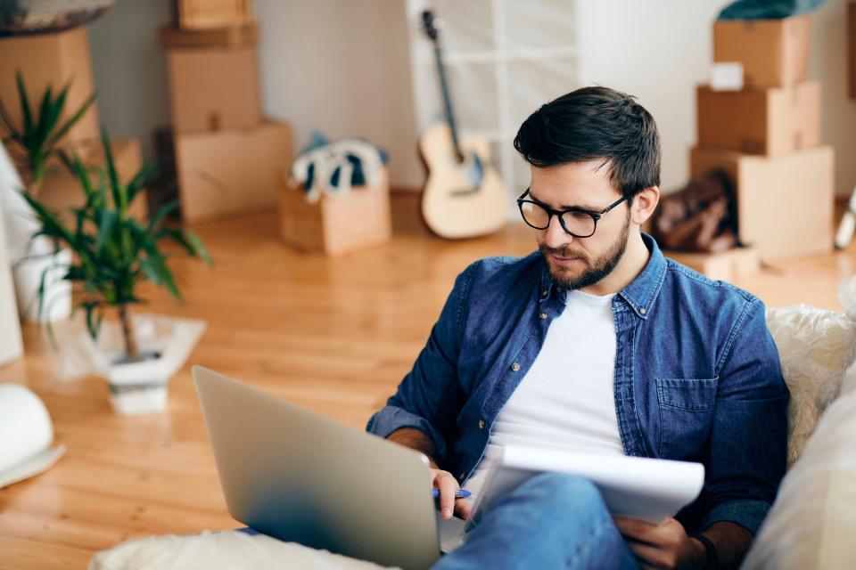 Man in blue shirt lays on a couch with a laptop and notepad in his lap.